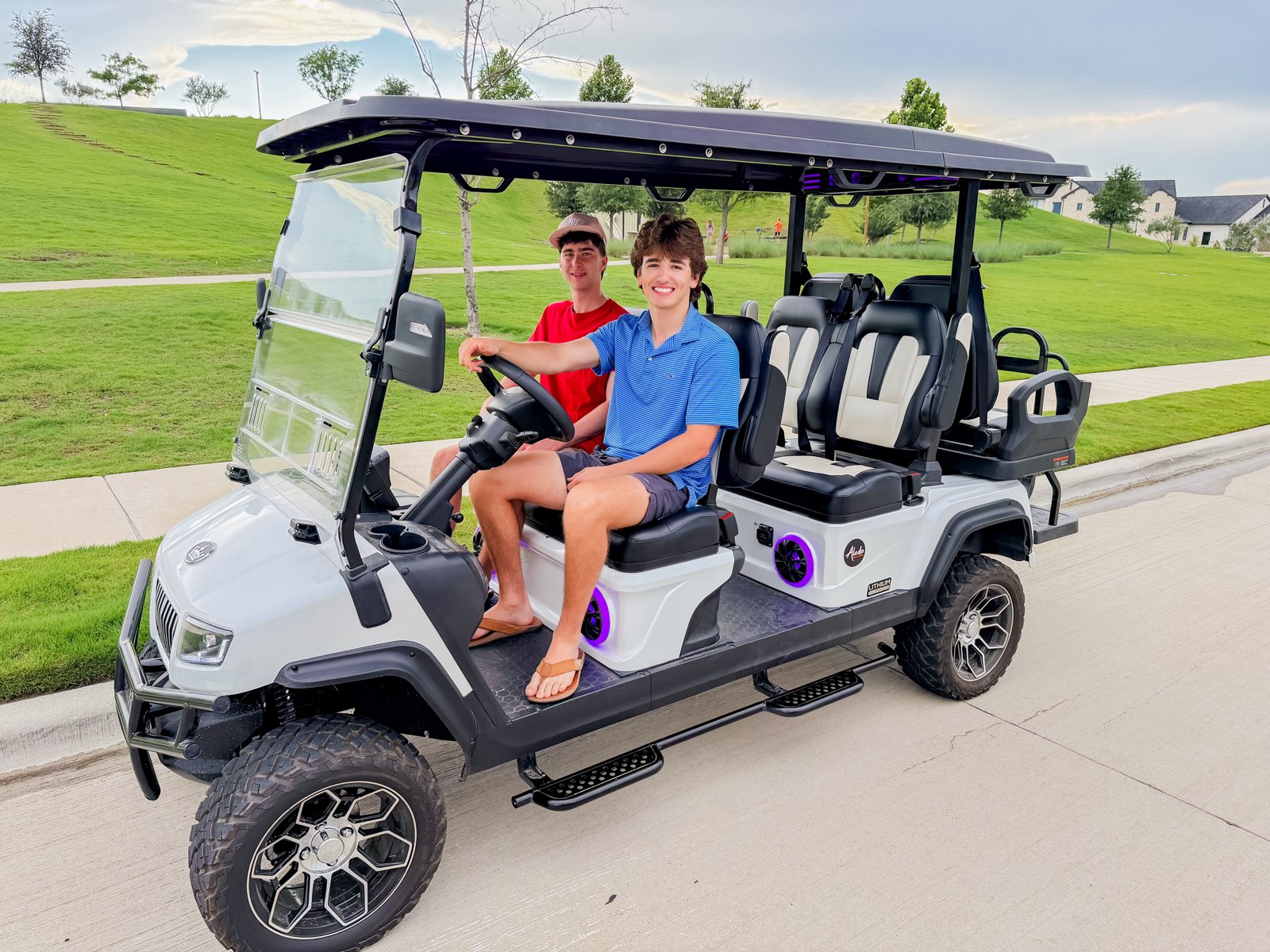 Two teenage boys sit in the front row of a white six-seat golf cart parked on a suburban street; the driver wears blue, the passenger wears red.